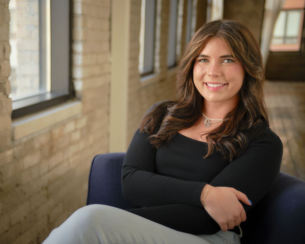 Professional headshot of someone in a black sweater sitting in a blue chair by a window with natural lighting.