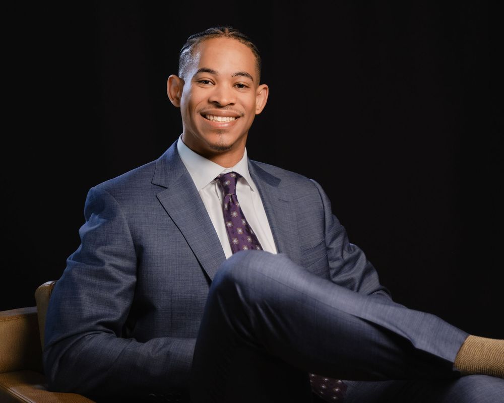 Professional wearing a dark suit and purple tie smiles while seated against a black background in a photo series.