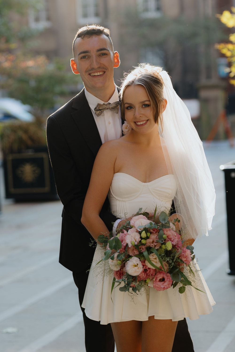 Newlyweds pose outdoors with bride holding pink and white bouquet in short wedding dress.