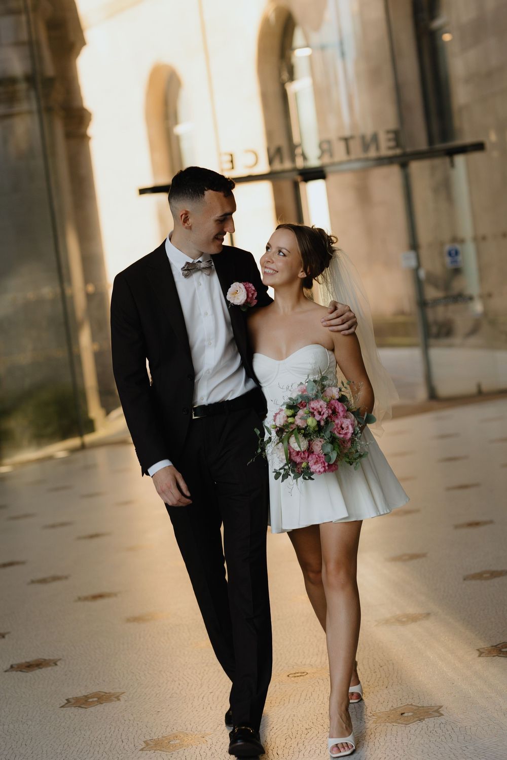 Newlyweds share intimate moments while walking together in warm sunlit hallway.