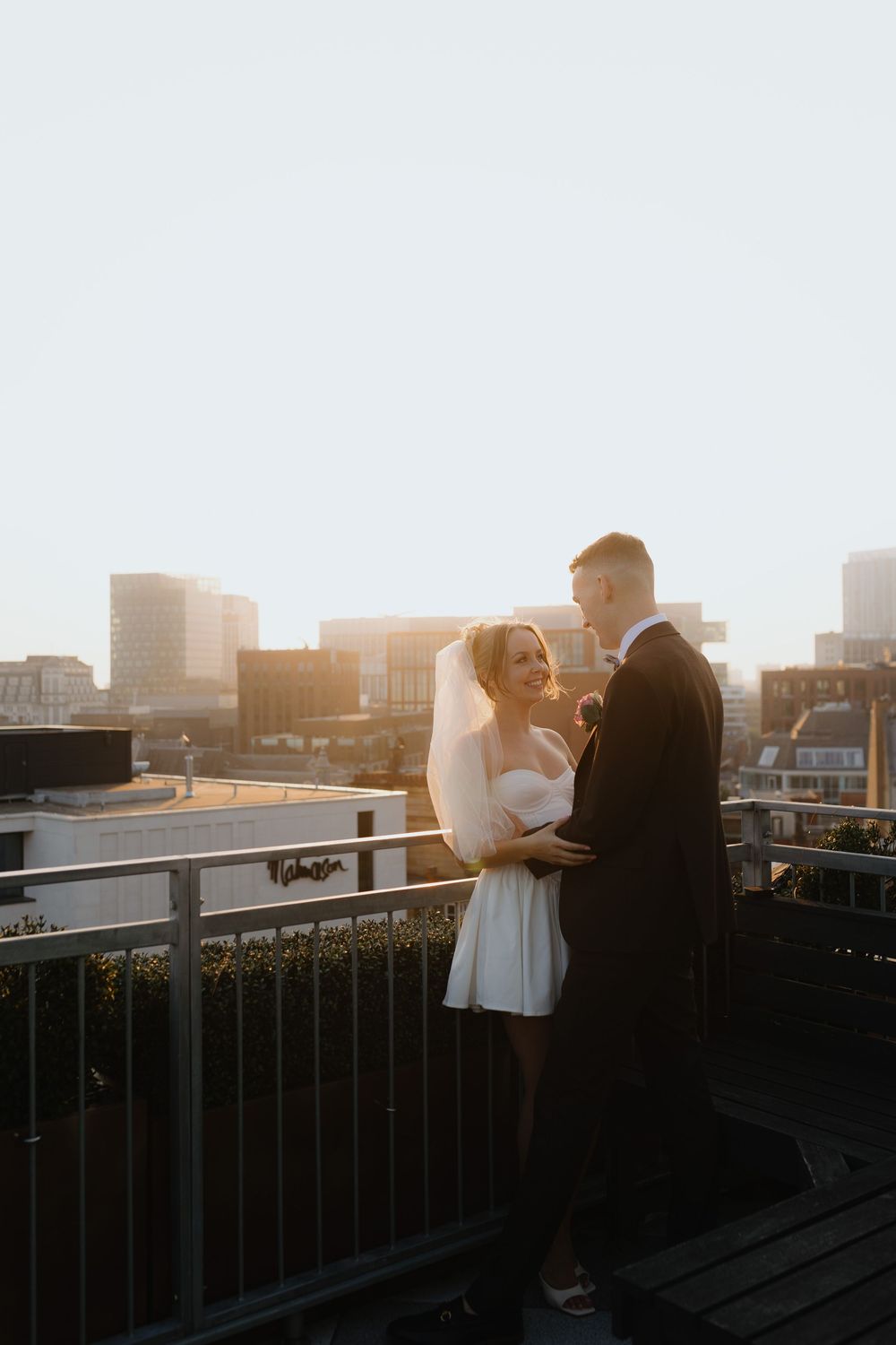 A couple shares an intimate moment on a rooftop at sunset with a cityscape backdrop.