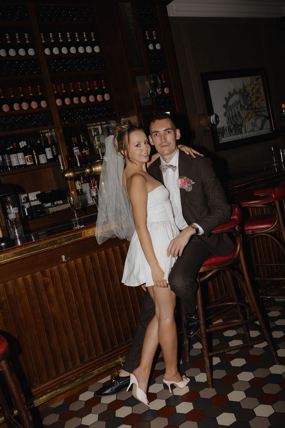 Newlywed couple posing at vintage bar with white dress and dark suit against wine bottle display.