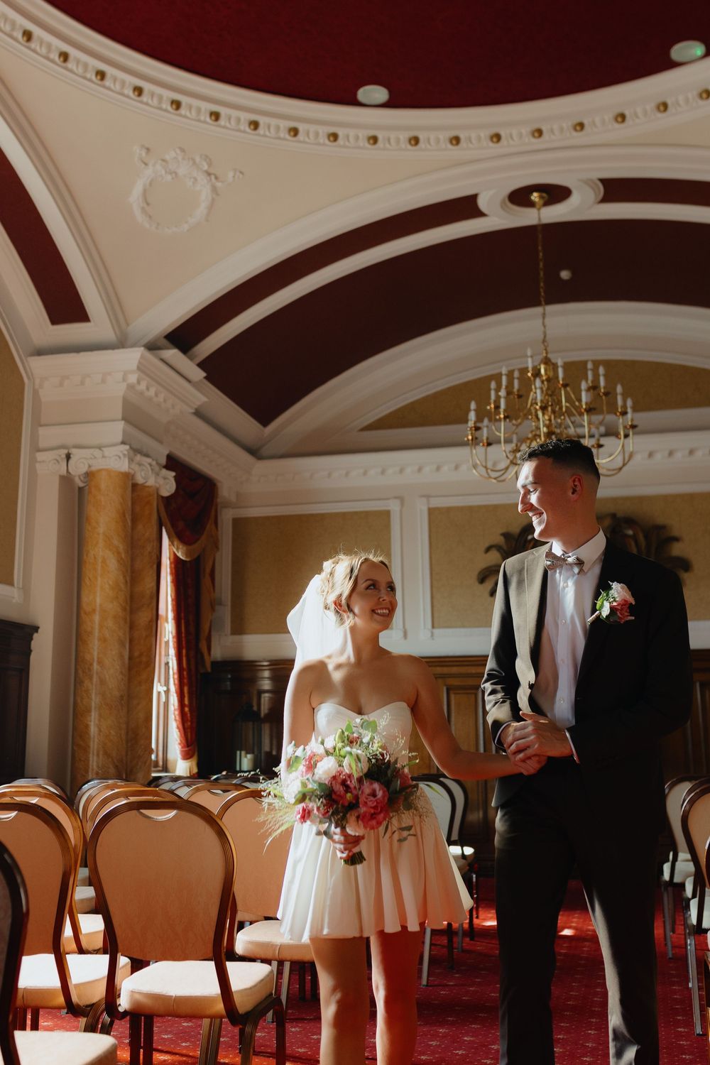 A wedding ceremony in an elegant room with ornate ceiling details, chandelier and red carpeting.