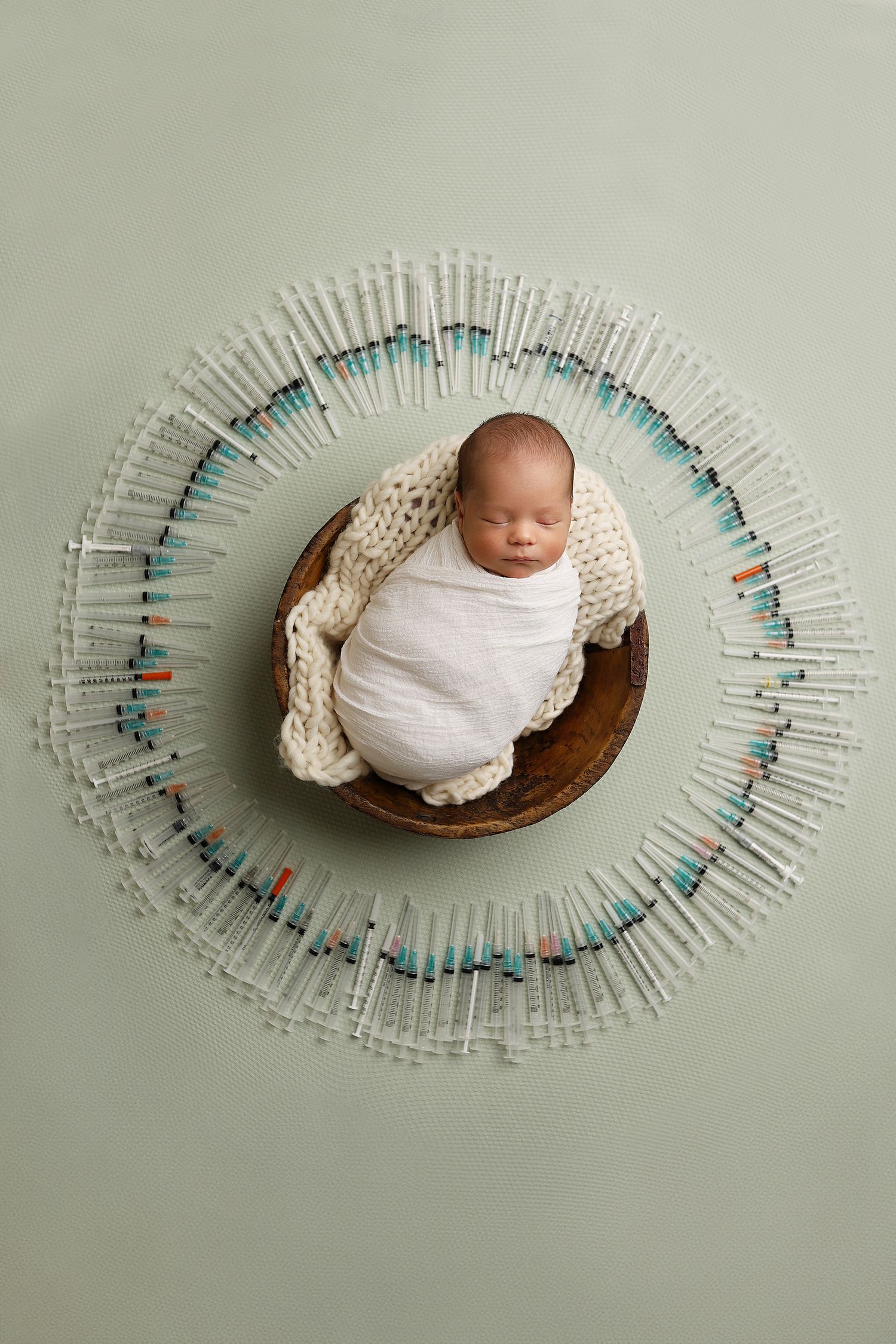 A swaddled baby lying in a wooden bowl surrounded by a circular arrangement of clothespins in a creative newborn photo. IVF baby photography