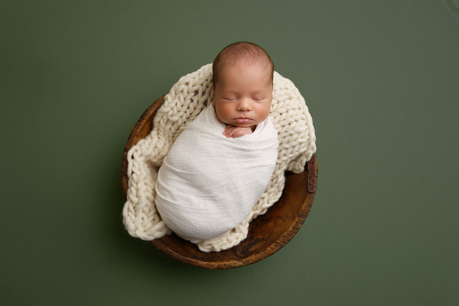 A newborn baby wrapped in white fabric rests in a wooden bowl against a dark green background. Orem newborn photography