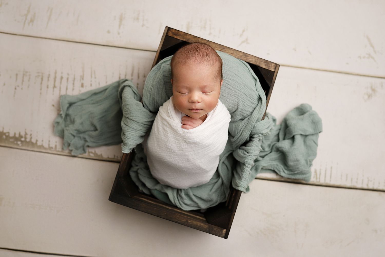 A series of newborn portraits showing a baby swaddled in white and sage green fabrics on wooden props. Utah County newborn studio