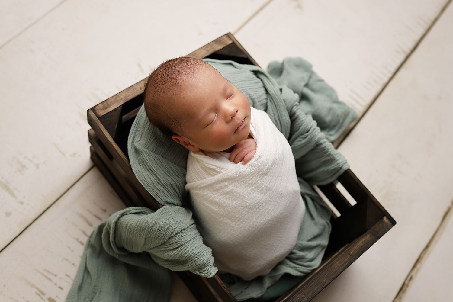 A newborn baby wearing a sage green cardigan and white onesie lies peacefully on a wooden surface. Orem newborn photographer