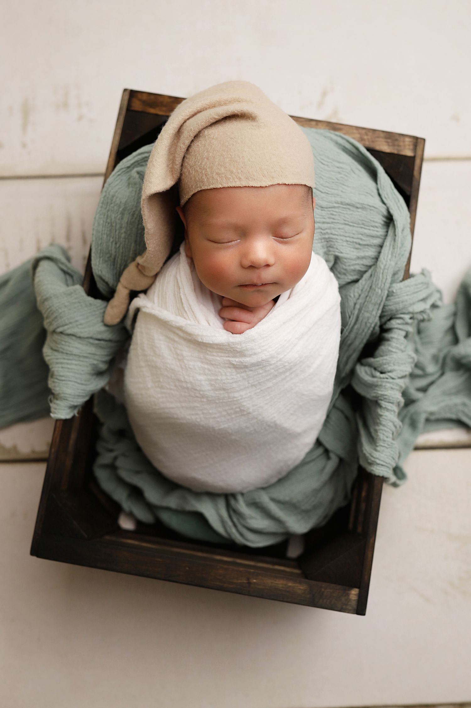 A sleeping newborn baby wrapped in white and sage green swaddle blankets rests peacefully in a wooden box. Utah County newborn photographer