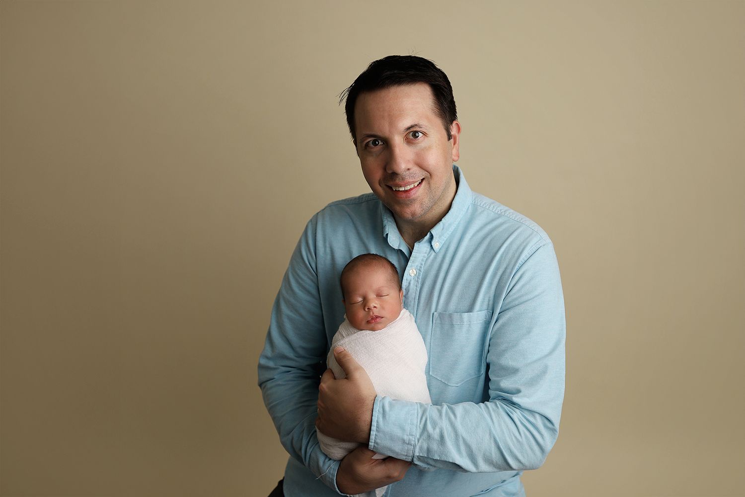 A parent in light blue shirt holds and gazes lovingly at a newborn baby in white against a beige background. Utah newborn photographer