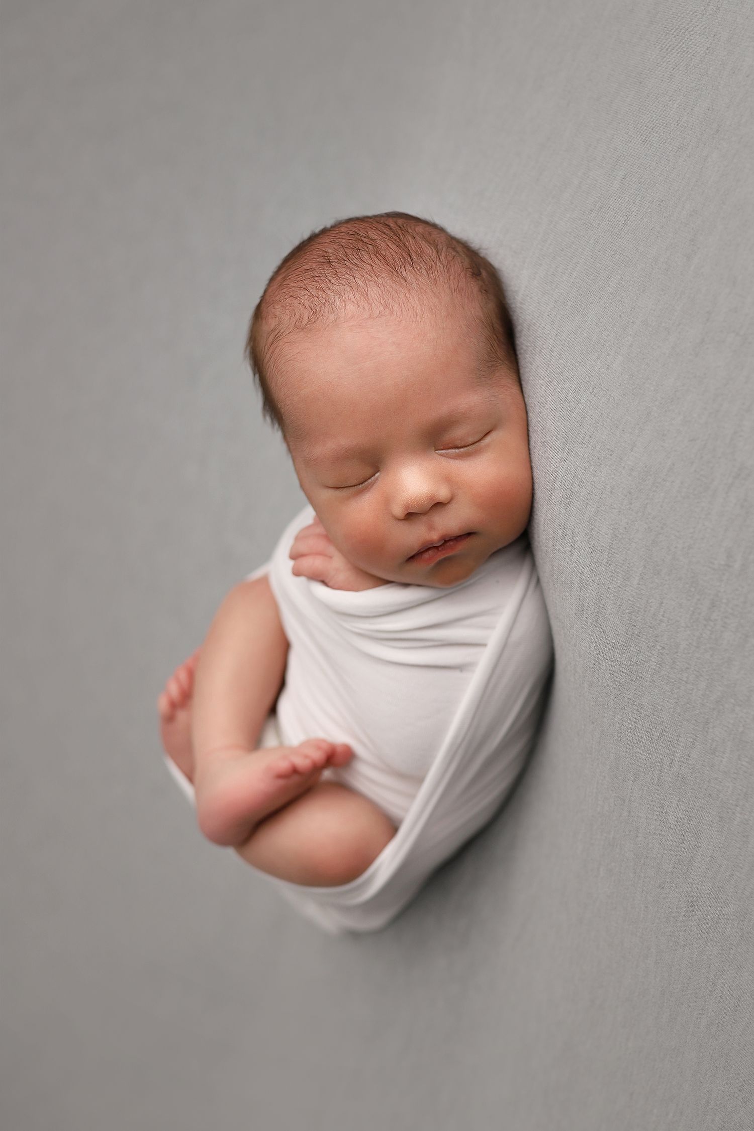 A sleeping newborn baby wrapped in white fabric against a gray background in a minimalist photography style.