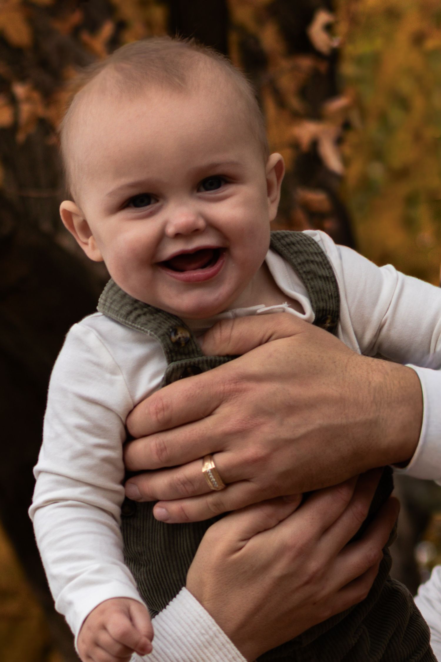 Laughing baby in green corduroy overalls held securely by adult hands with gold ring.