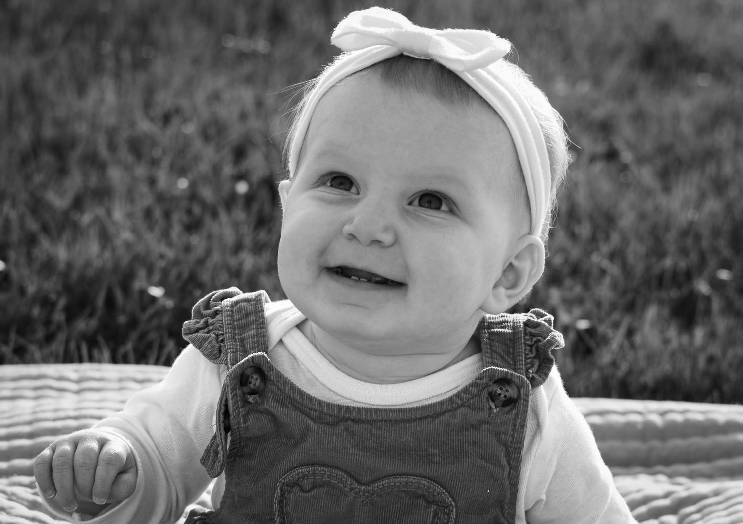 Black and white portrait of a smiling baby girl wearing a bow headband and overalls.