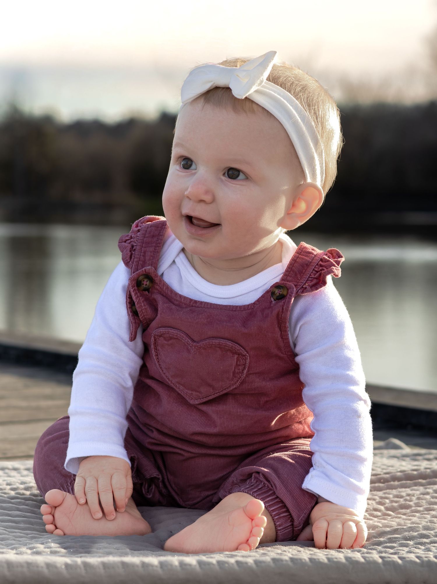 Adorable baby girl in pink overalls and white bow headband sitting on a dock.
