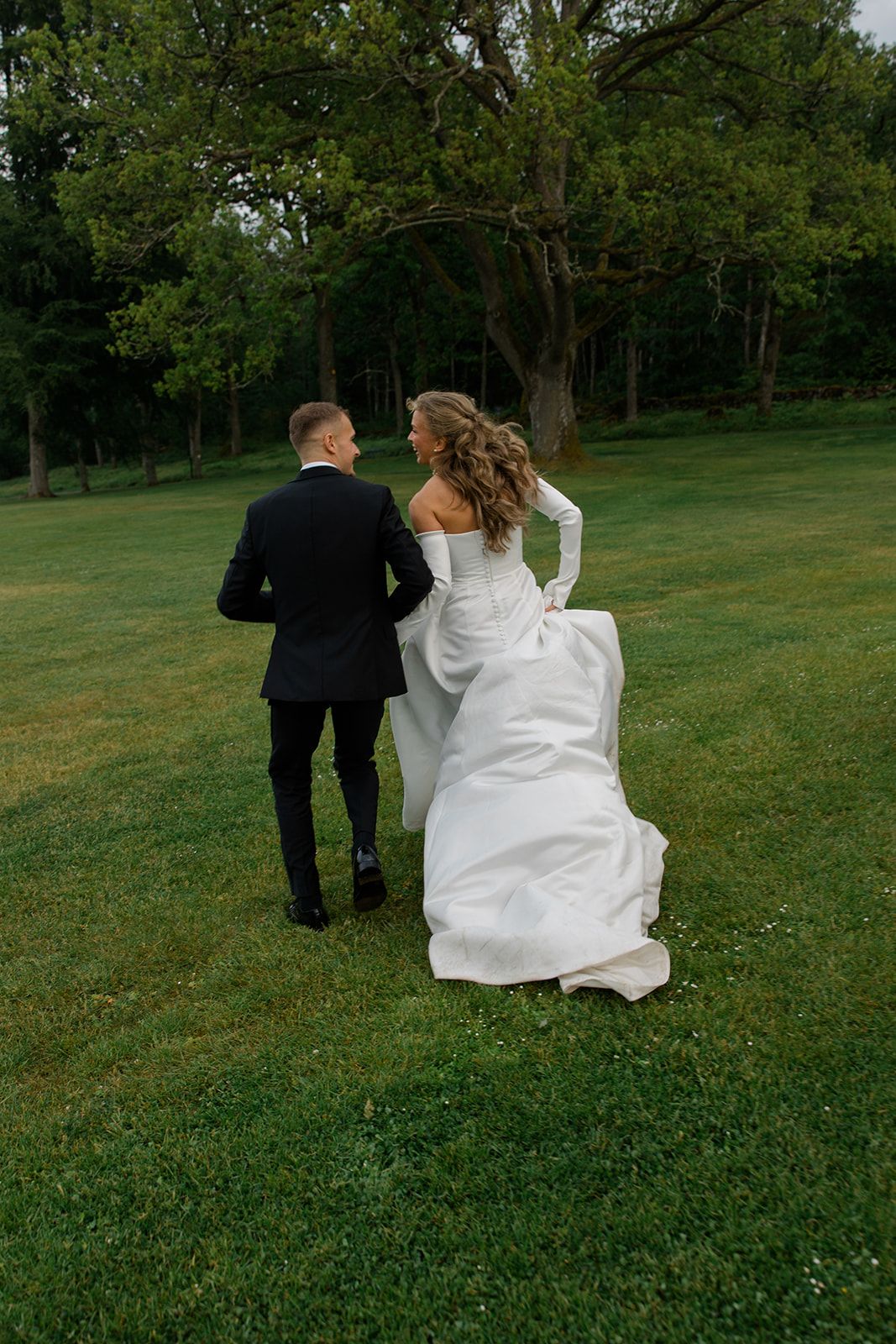 A couple in wedding attire runs playfully through a lush green field on their special day.