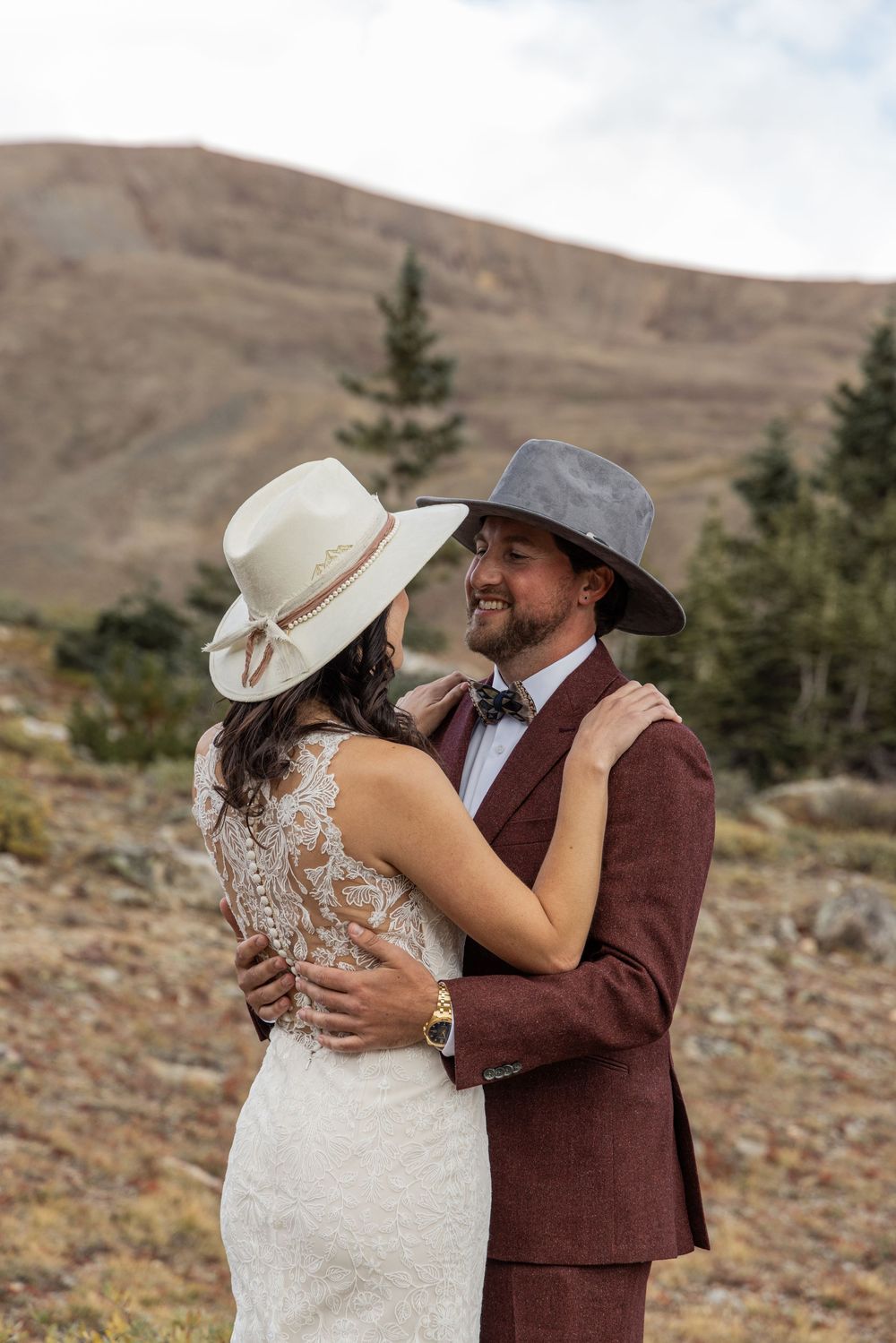 A couple in western attire embrace during a mountain wedding photoshoot, wearing cowboy hats against a rugged landscape.