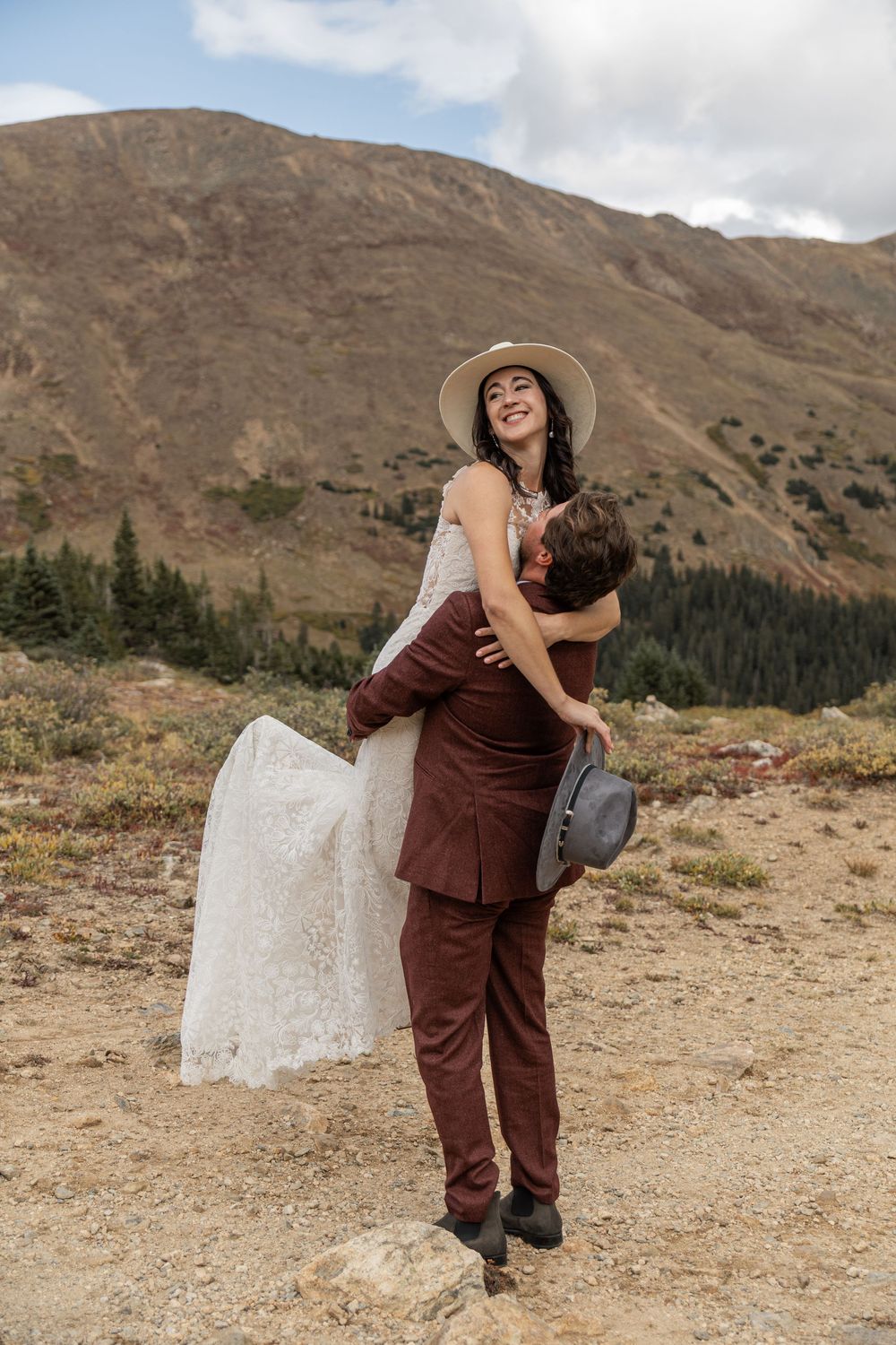 A couple embraces in matching brown outfits against a scenic mountain backdrop in the wilderness.