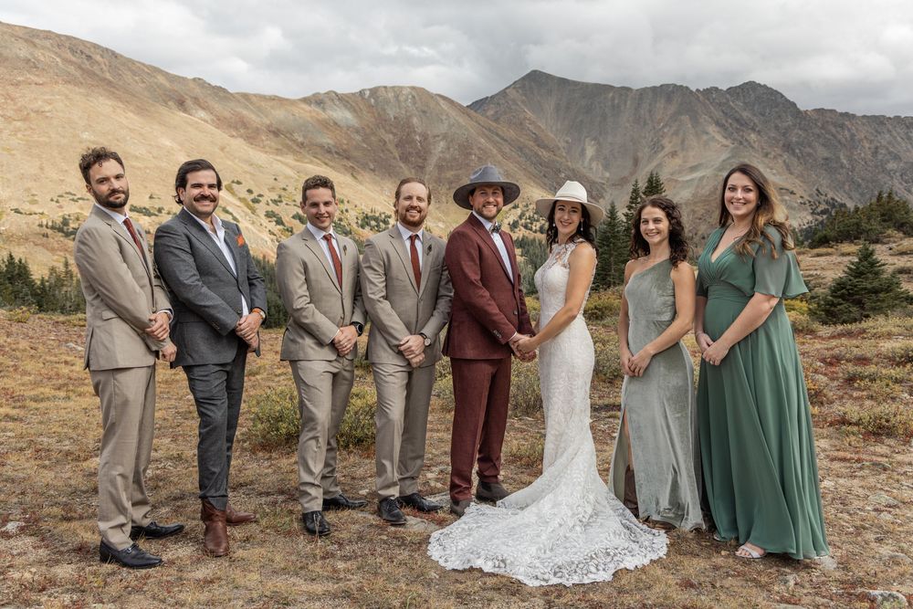 A wedding party poses together against a backdrop of rugged mountain peaks in a desert landscape.