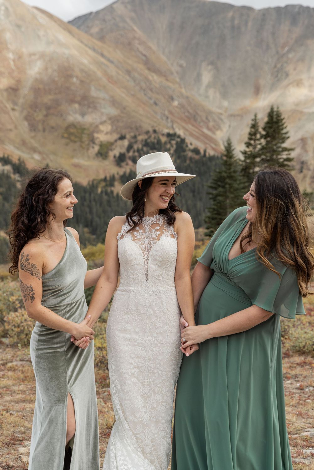 A group in sage green dresses and white lace share joyful moments against a mountainous backdrop.