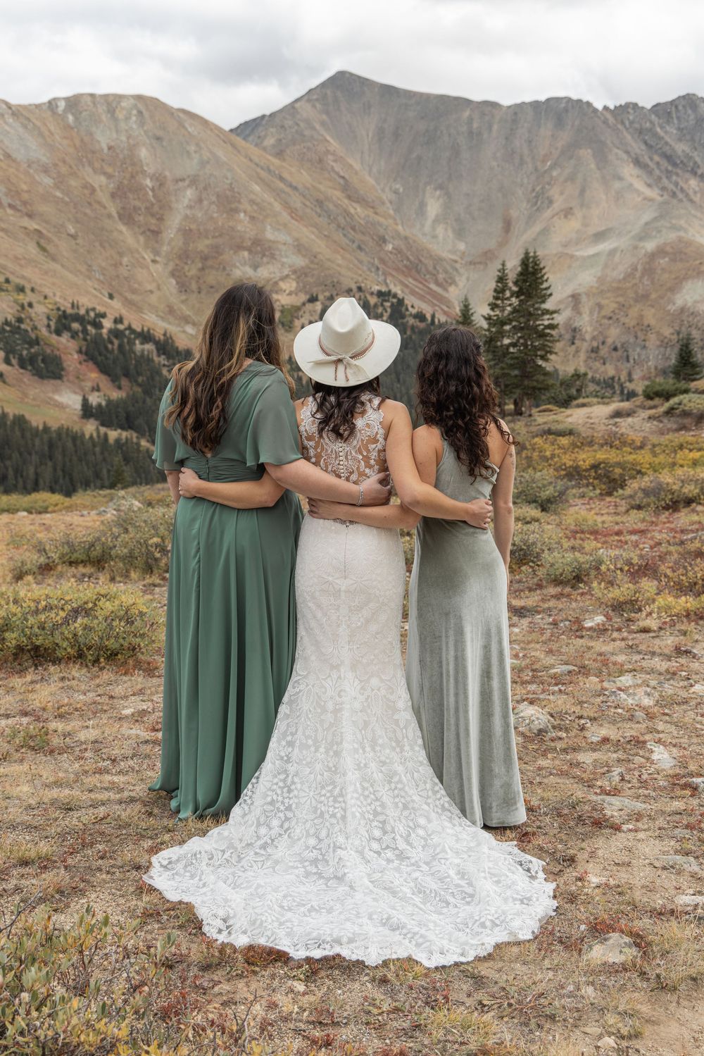 Three figures in green and white dresses stand together against a backdrop of majestic mountain peaks and autumn terrain.