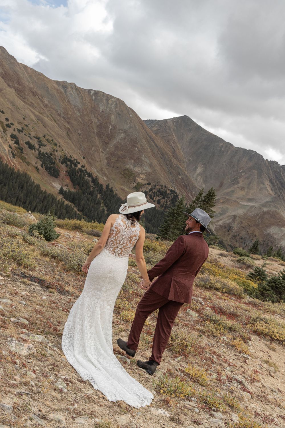 A couple in wedding attire walks up a rocky mountain slope with dramatic cloudy skies and peaks in the background.