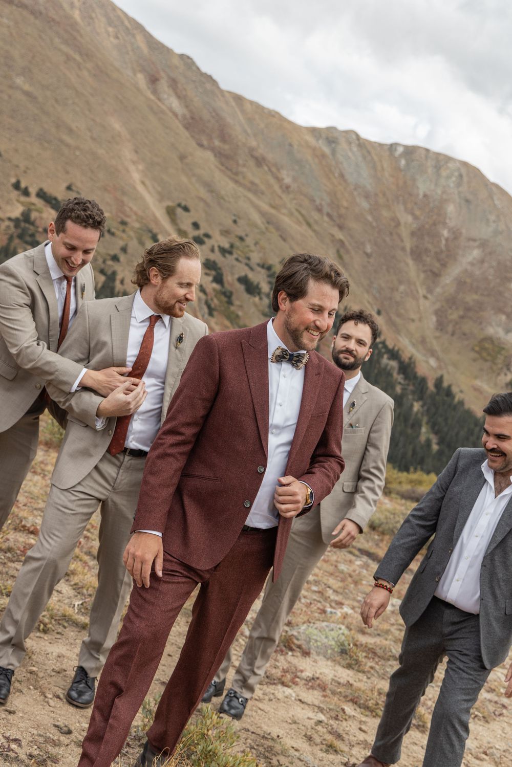 A group of groomsmen in suits playfully pose together against a mountainous backdrop in the wilderness.
