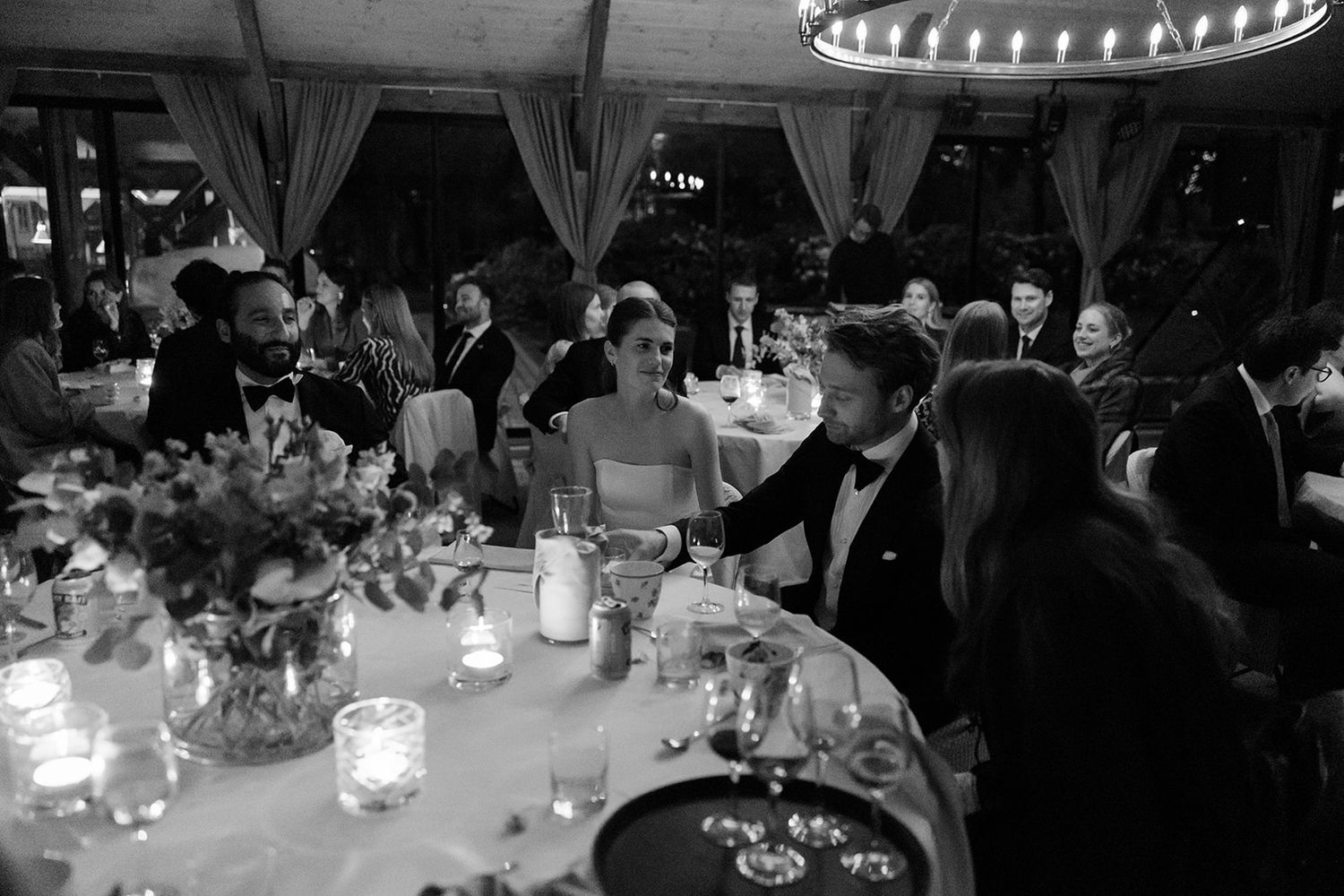 Elegant black and white reception scene with candlelit tables, floral centerpieces and guests gathered under circular lighting.