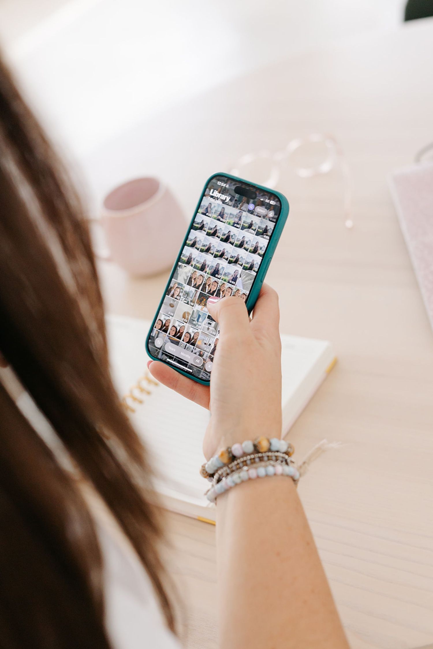 Hand holding a smartphone displaying a photo grid interface with teal-colored case and bracelet accessory.