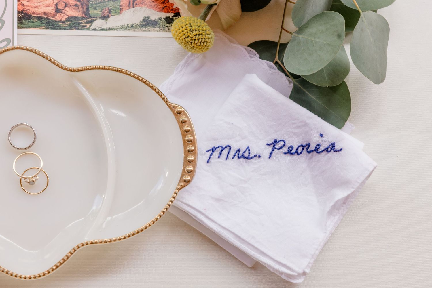 Delicate white monogrammed handkerchief with embroidered scalloped edges displayed with eucalyptus leaves and gold jewelry.
