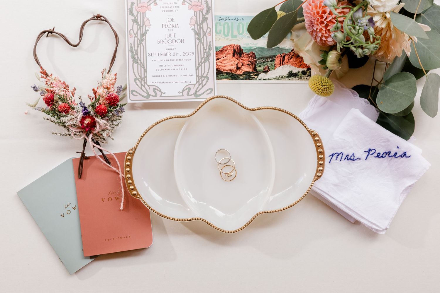 Wedding details flatlay with floral heart decoration, rings on white plates, and eucalyptus leaves with coral flowers.