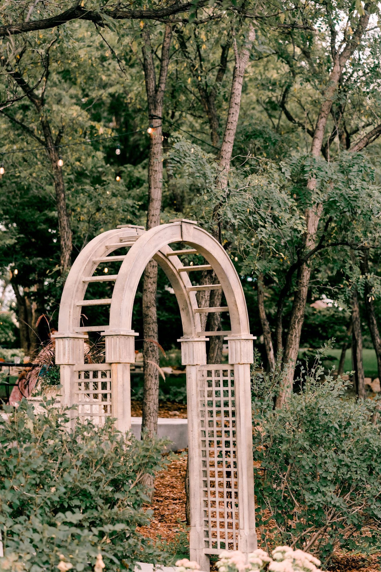 A wooden garden arbor with lattice sides stands in a peaceful wooded setting surrounded by trees and foliage.