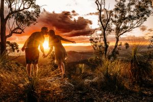 Two silhouettes embrace during a dramatic mountain sunset with sunlight filtering through trees.