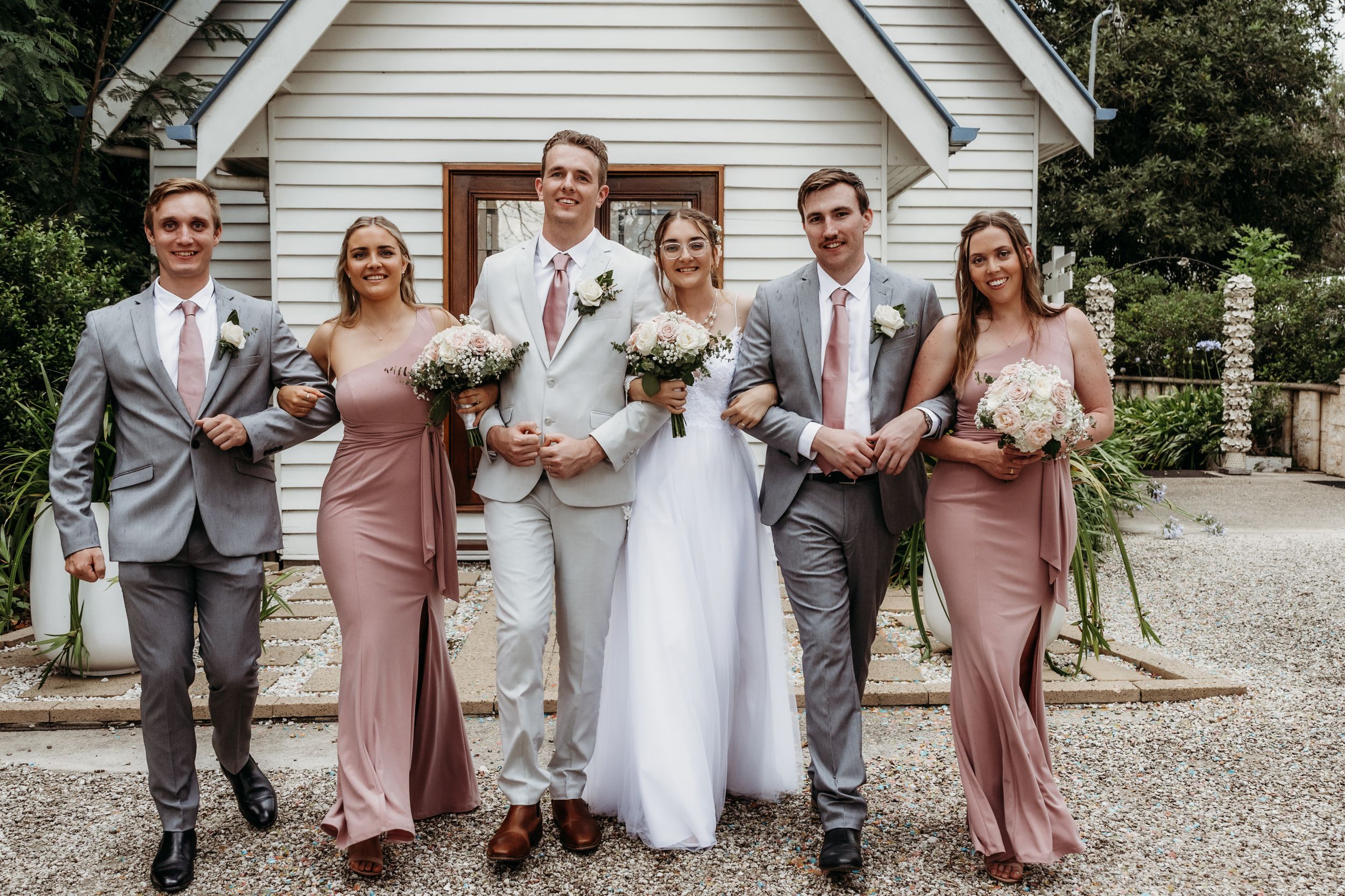 Wedding party in gray suits and pink dresses posed in front of white chapel.