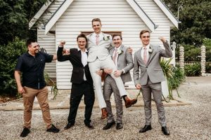 Groomsmen lifting groom in celebratory pose outside white chapel.