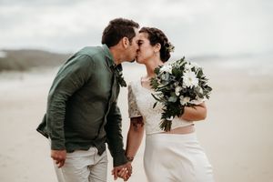 A romantic couple shares an intimate kiss on a misty beach while holding a white and green floral bouquet.
