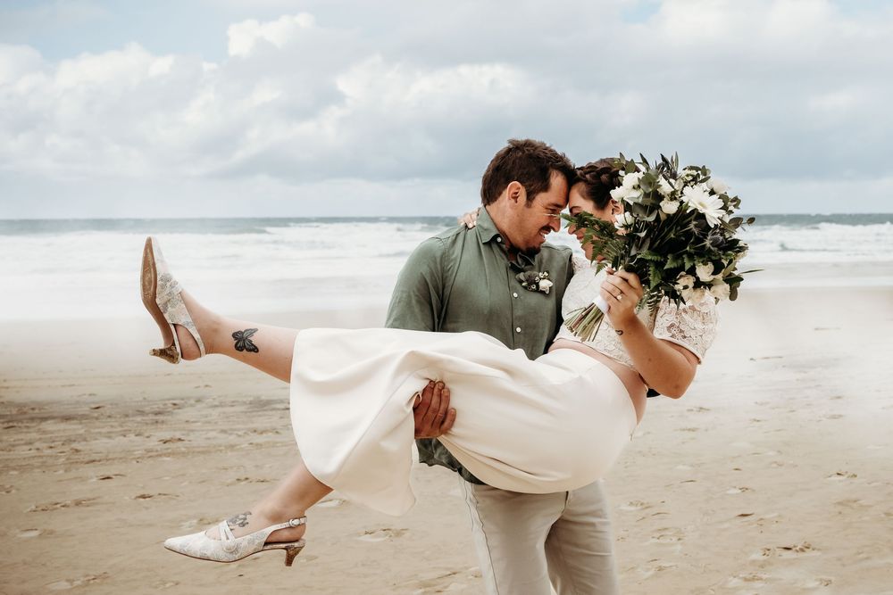 A couple shares a joyful moment on a cloudy beach as they embrace with a white floral bouquet.