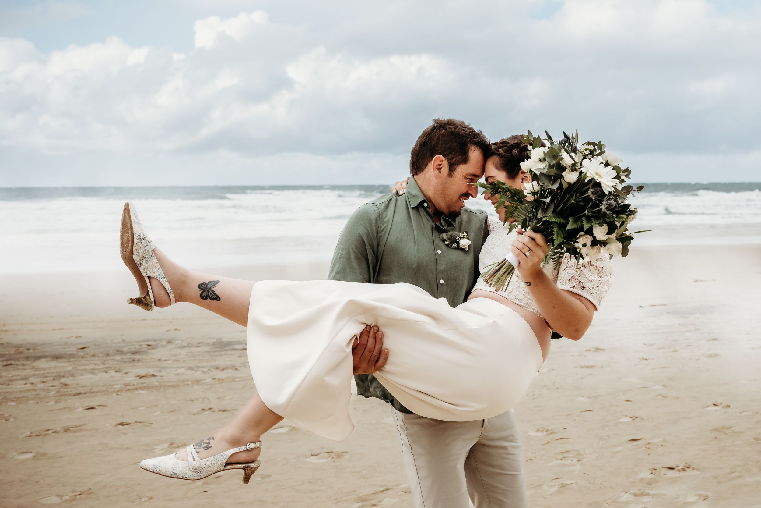 A couple shares a joyful moment on a cloudy beach as they embrace with a white floral bouquet.