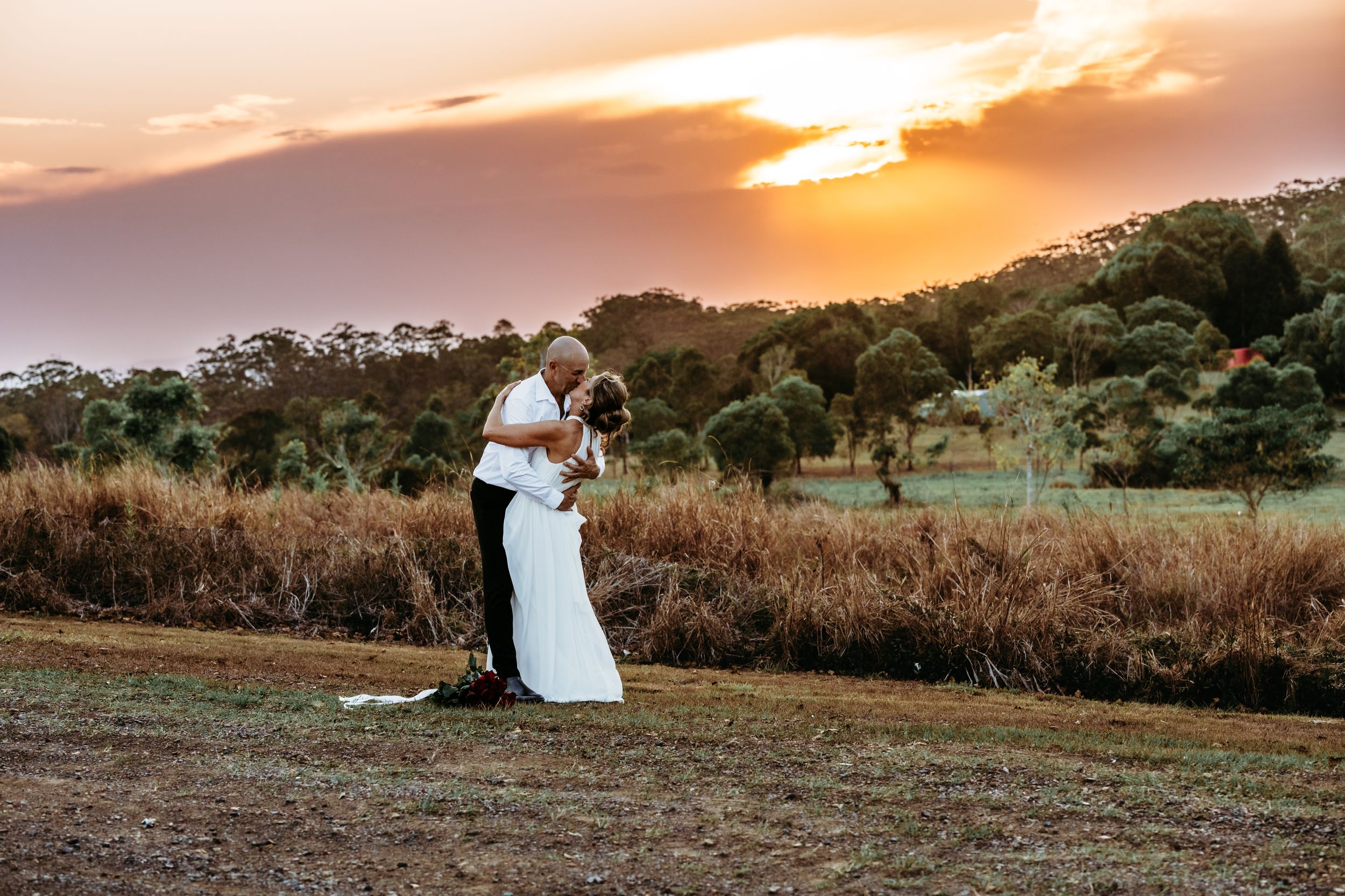 Silhouetted couple embraces in an open field during a golden sunset with tall grass and trees in background.