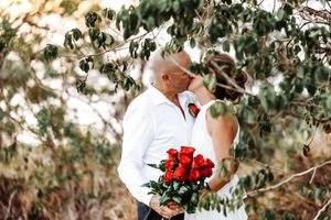 A romantic couple embraces under lush green trees while holding red roses in a garden setting.