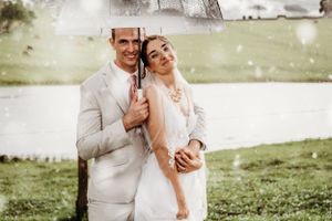 A joyful wedding couple laughs together outdoors in formal white attire on a grassy hillside.