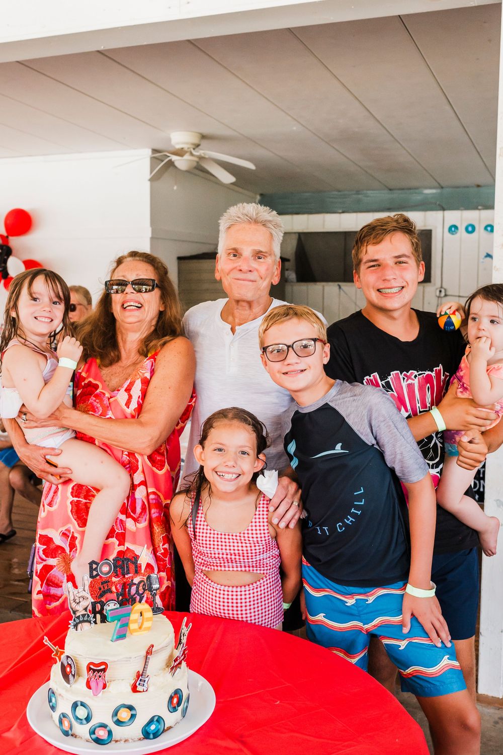A family gathering around a birthday cake with red and white decorations on a table.