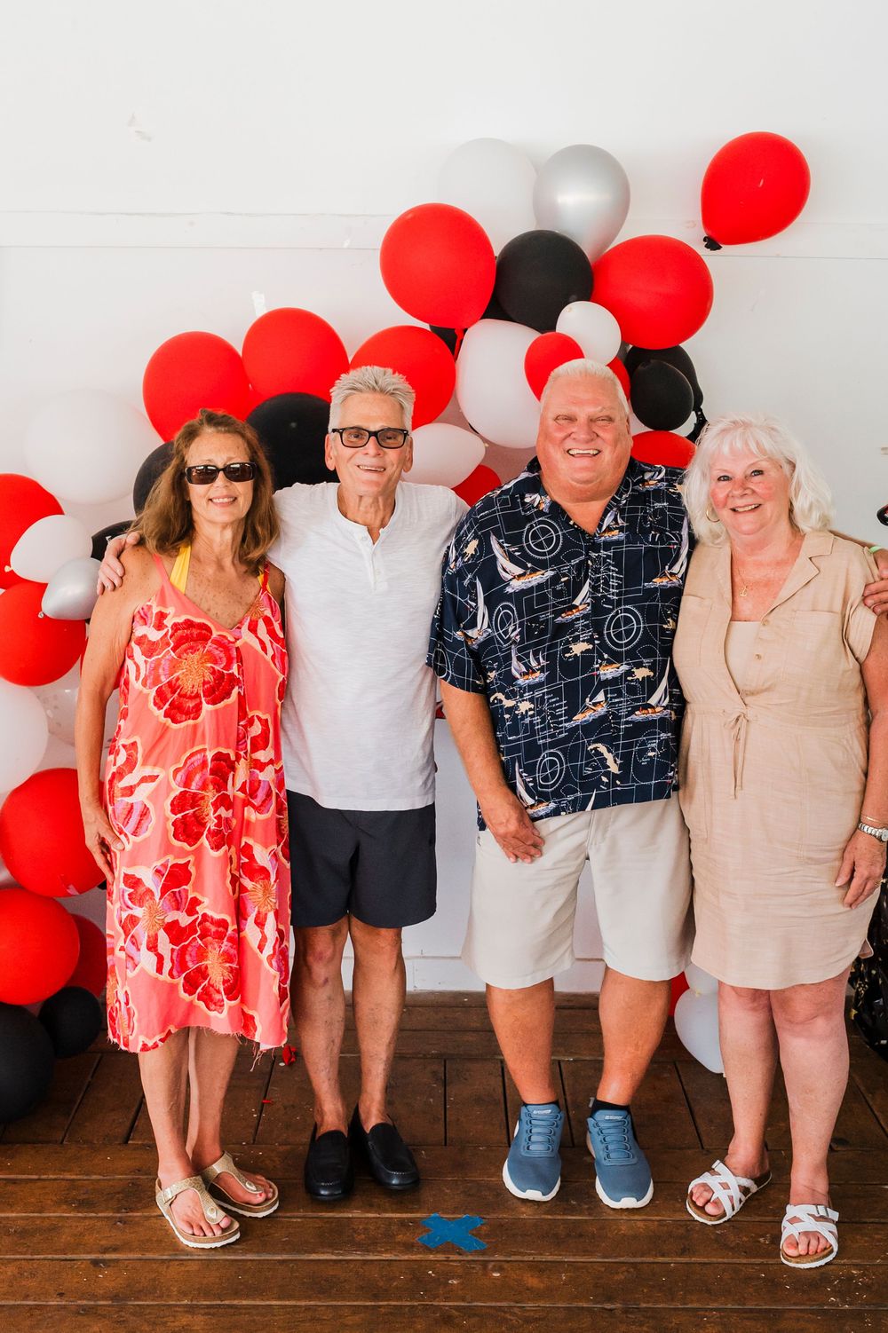 A group photo with red and black balloons against a white wall, showing people in summer attire.
