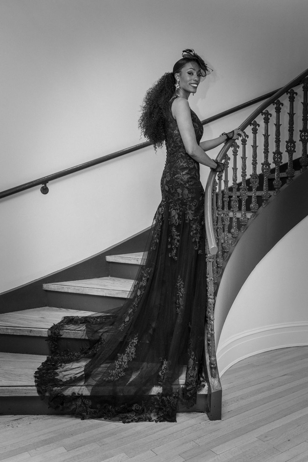 A flowing black lace gown captured in motion along an elegant staircase with white walls and wooden banister.