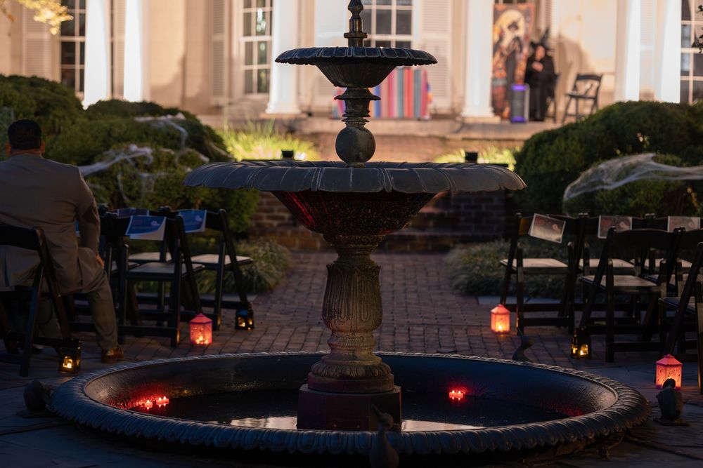 An elegant fountain illuminated by red lights at dusk with outdoor seating and a historic building in the background.