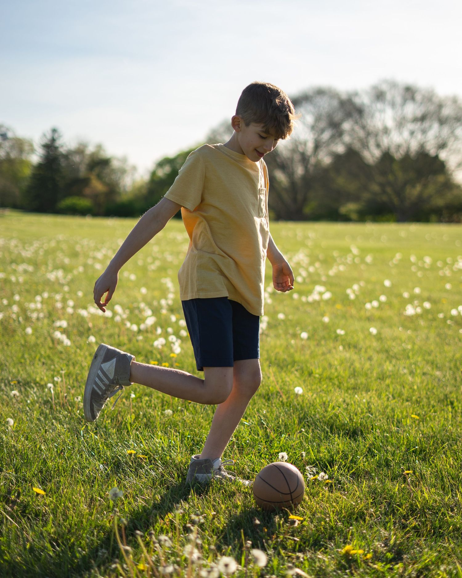 A young boy in a yellow shirt and blue shorts kicks a basketball on a sunny green field dotted with dandelions.