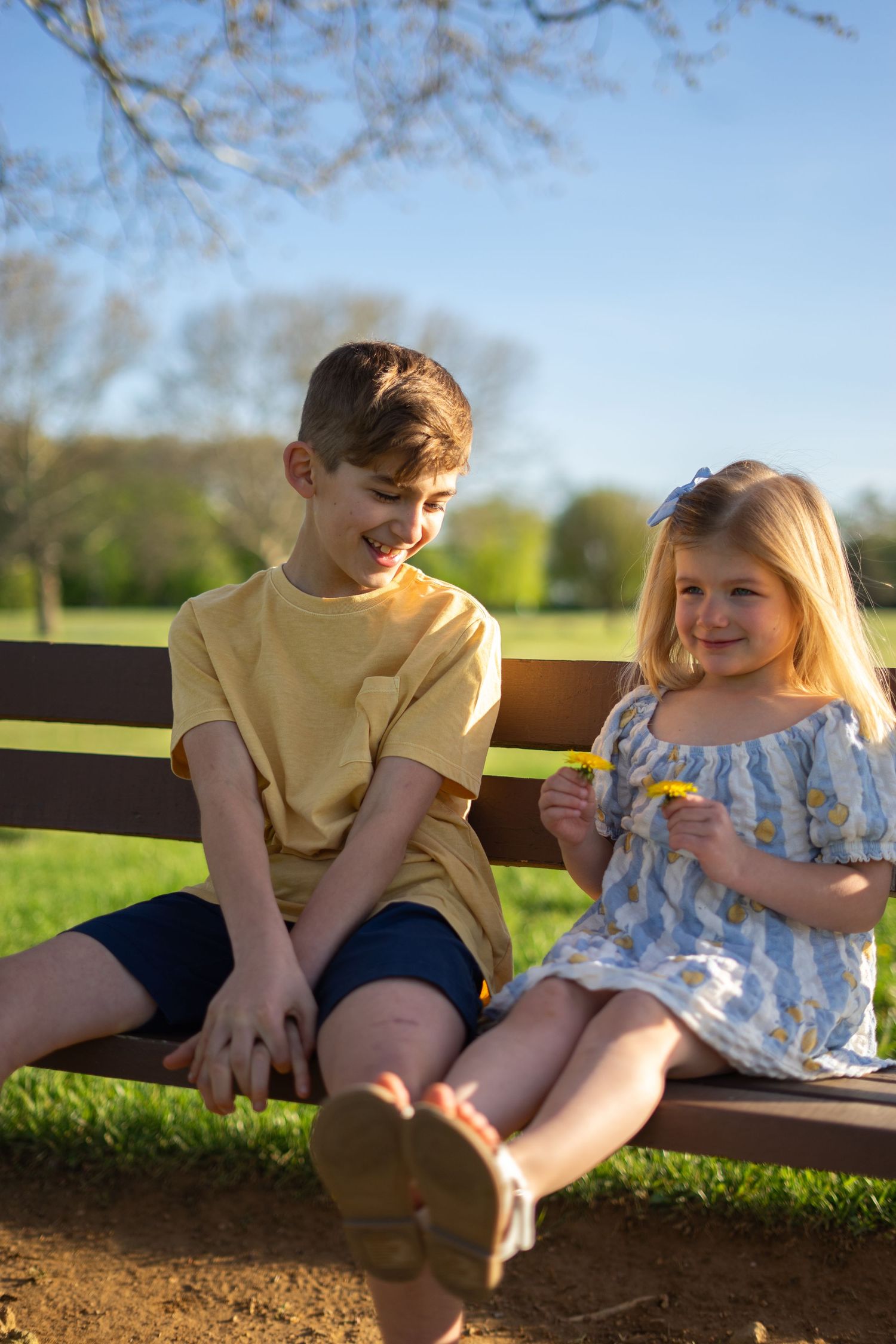 Two children sitting on a park bench outdoors on a sunny day, boy in yellow shirt smiling at girl in floral dress.