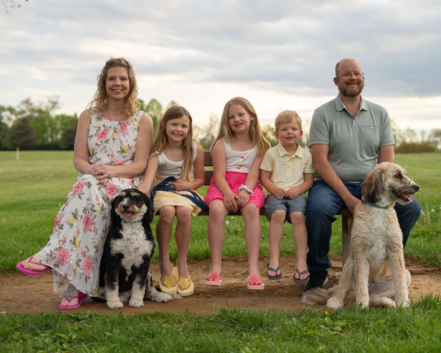 Family of five with two dogs sitting on a bench outdoors in a green park setting for a portrait.
