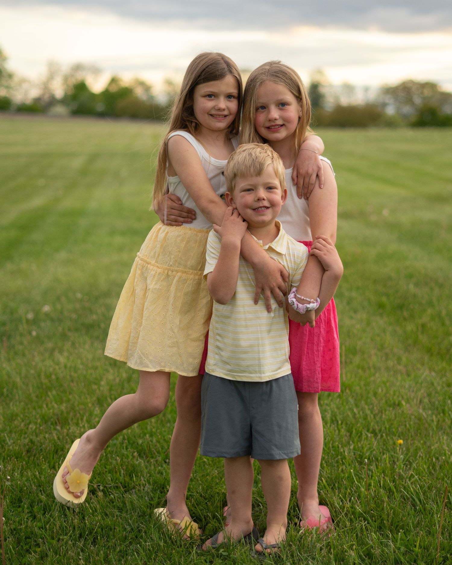 Three children hugging and smiling together in a green field at sunset, wearing summer outfits.