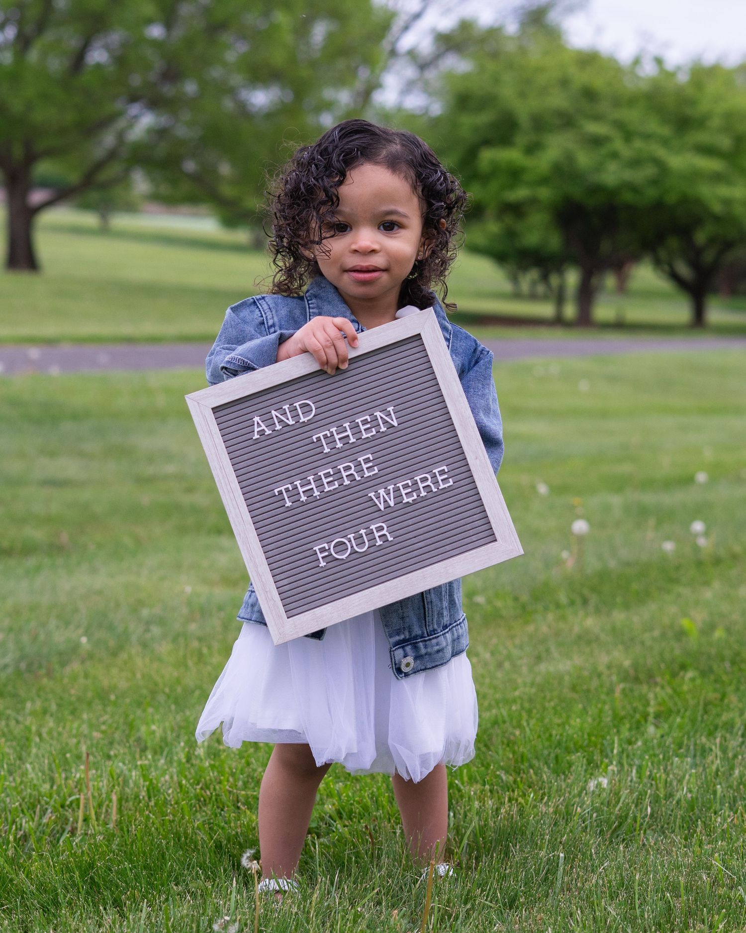 Toddler girl in denim jacket holds letter board sign reading And Then There Were Four on green grass.
