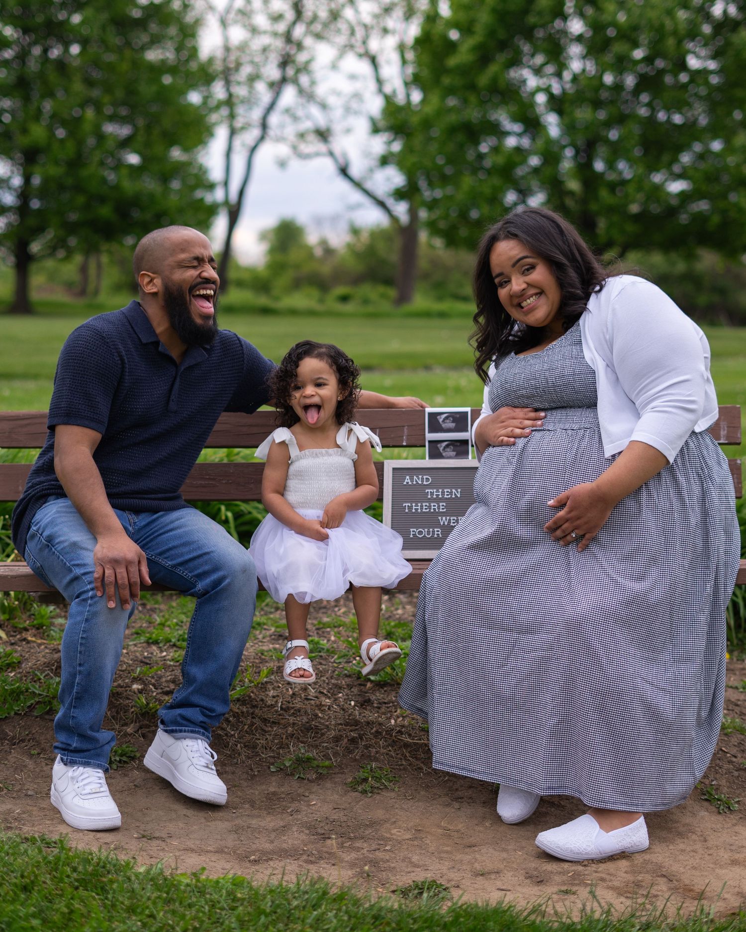 Happy family maternity photo shoot in park with laughing dad, excited toddler girl, and pregnant mom holding belly.