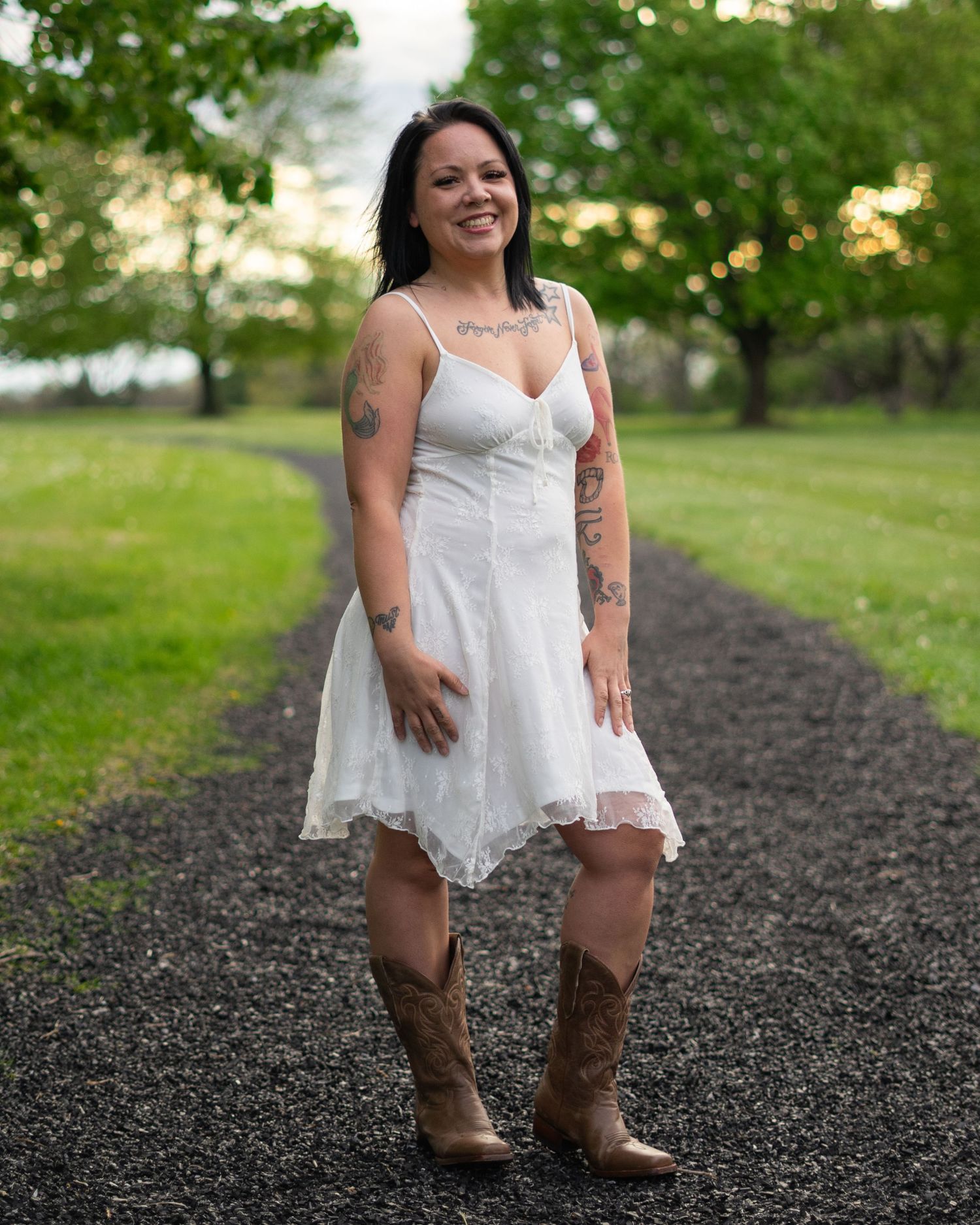 Woman in white sundress and cowboy boots smiling on a gravel path surrounded by lush green trees.